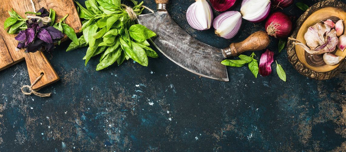 Healthy food cooking background. Fresh green and purple basil leaves, red onions and garlic with herb chopper knife and rustic cutting board over grunge dark blue plywood texture. Top view, copy space